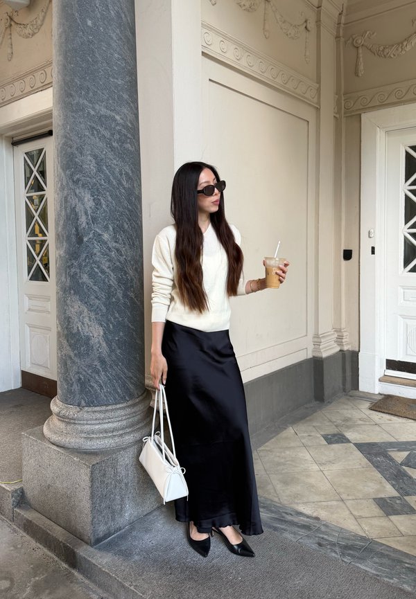 Femme aux longs cheveux foncés portant des lunettes de soleil, un pull blanc, une jupe noire et des talons, tenant un sac blanc et un café glacé, debout près d'une colonne en marbre.