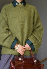 Olivově zelený chlupatý svetr s tmavě zeleným límcem, zlaté šperky s perlami, šedá látková sukně a hnědá kožená kabelka.