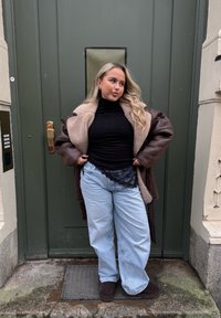 Jeune femme aux longs cheveux blonds portant un col roulé noir, un manteau en peau retournée marron, un jean bleu clair et des chaussures foncées, debout devant une porte verte.