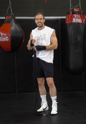 Boxeador con una camiseta sin mangas blanca con el texto "YALE", pantalones cortos negros y zapatillas altas blancas, envolviendo sus manos con cinta negra, de pie junto a un saco de boxeo rojo y negro.
