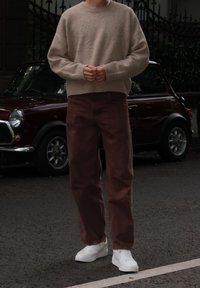 Beige uldtrøje, brune fløjlsbukser og hvide sneakers. Urban baggrund med en vintage bil delvist synlig. Teksturerede materialer.