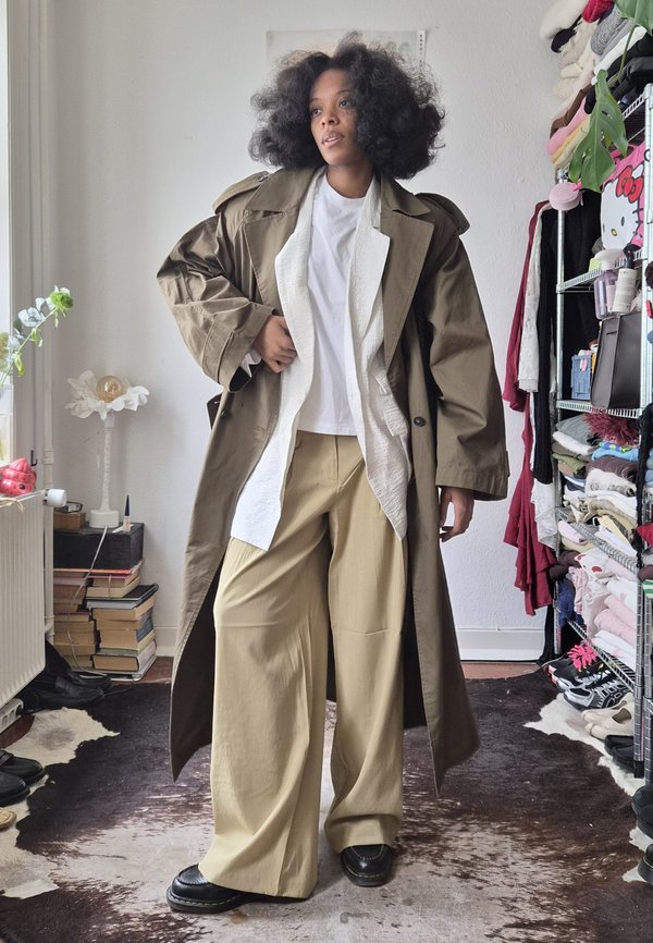 Woman with curly hair wearing a long brown coat, white shirt, beige wide-leg pants, and black shoes standing in a room with shelves and stacked books.