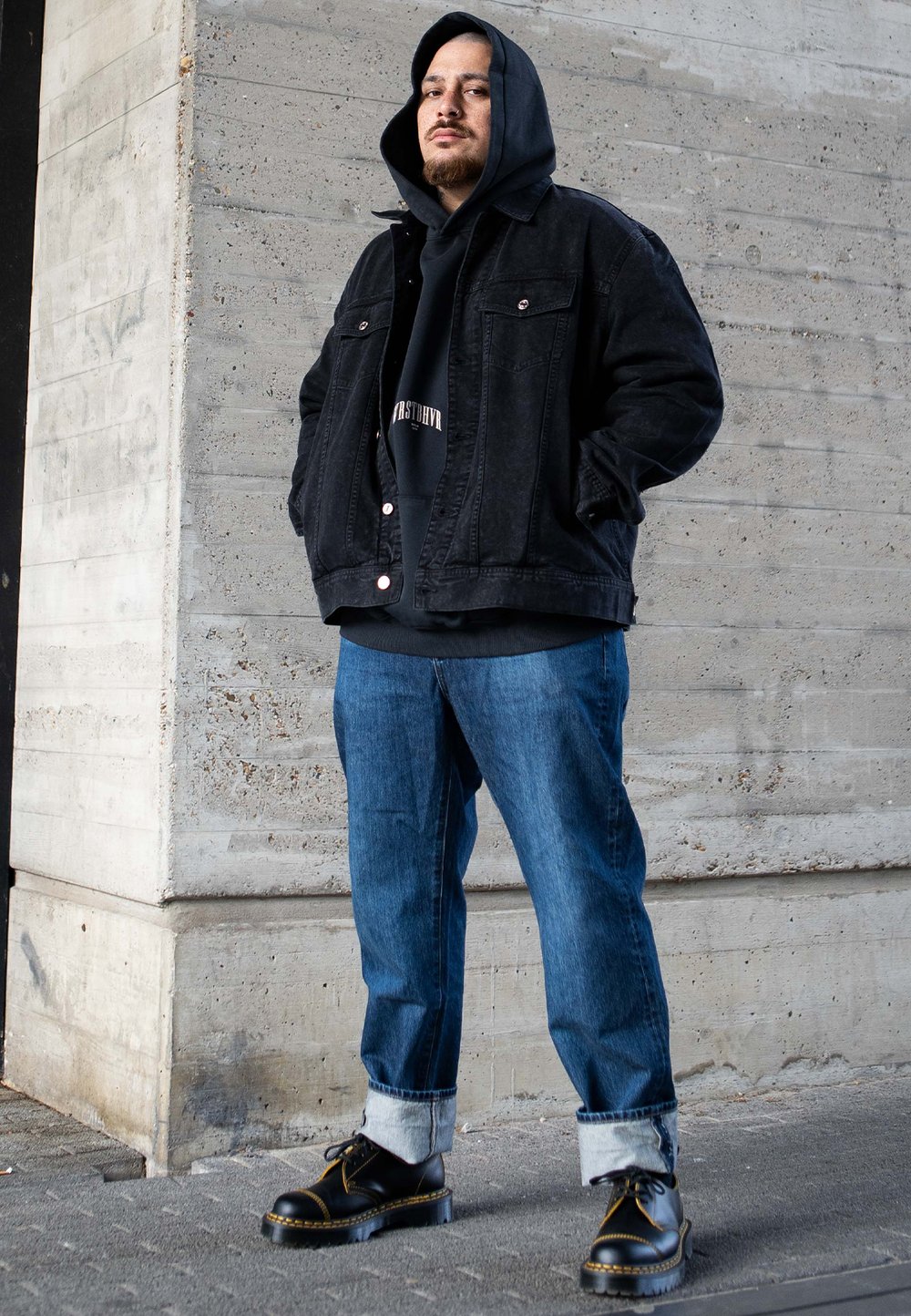 Fekete farmerdzseki fekete kapucnis pulóver felett, kék farmer tüskés szegéllyel, és fekete cipő sárga kiemelésekkel, egy beton háttér előtt.