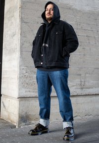 Fekete farmerdzseki fekete kapucnis pulóver felett, kék farmer tüskés szegéllyel, és fekete cipő sárga kiemelésekkel, egy beton háttér előtt.