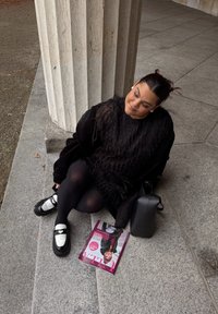 Černý oversize svetr s pírkovou texturou, černé legíny a boty s bílými akcenty. Na zemi černá taška a časopis.