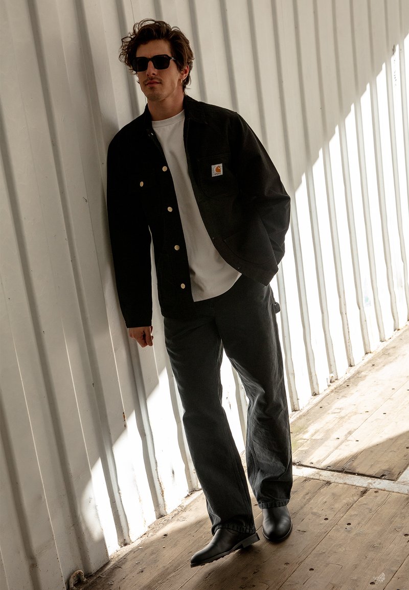 Juodas švarkas su auksinėmis detalėmis, balta marškinėliai, tamsūs džinsai ir juodi batai. Stovi prieš tekstūruotą baltą sieną su mediniais grindimis.