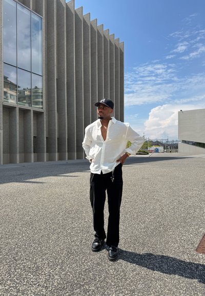 Camisa blanca de manga larga, desabrochada, combinada con pantalones negros y zapatos negros. De pie al aire libre, con un edificio moderno de fondo.
