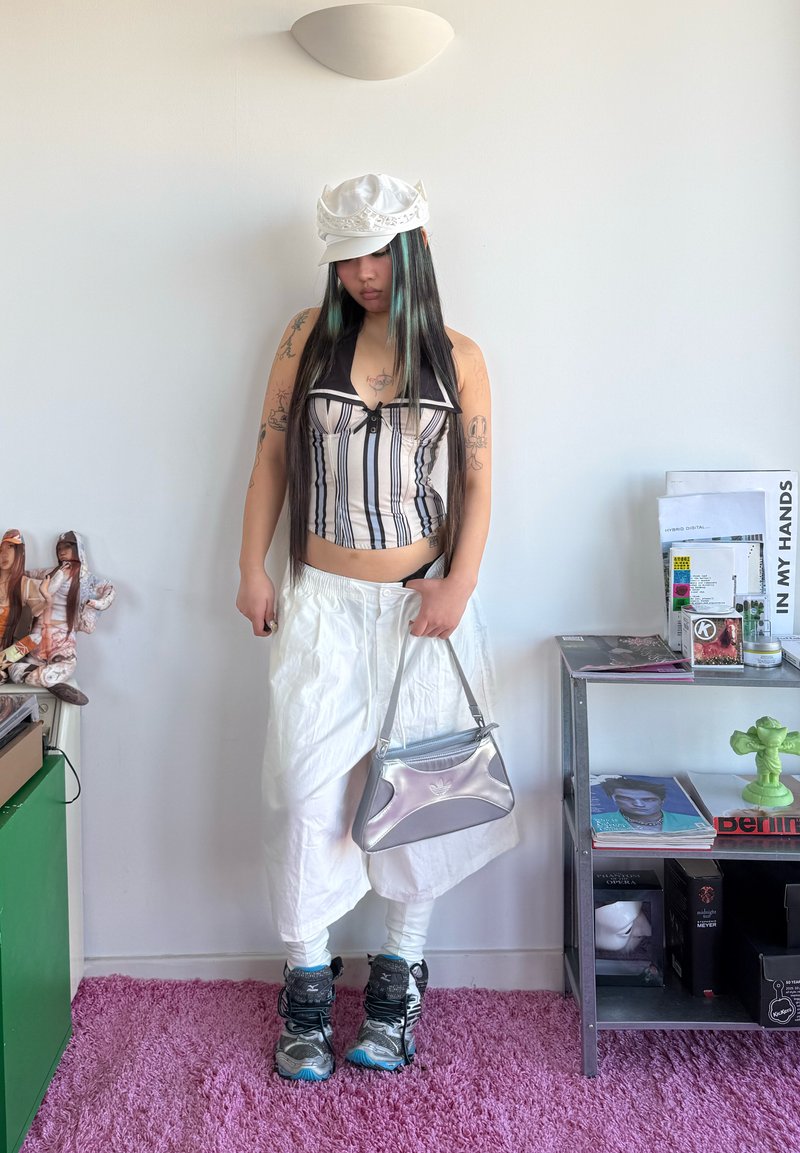 Tânăr cu păr lung, purtând o pălărie albă, top fără mâneci cu dungi, pantaloni largi albi, geantă argintie și adidași, stând în interior.