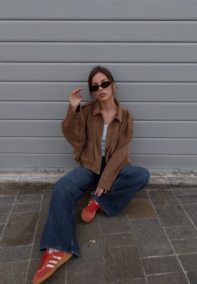 Mujer con chaqueta marrón, vaqueros azules de pierna ancha, zapatillas rojas y gafas de sol negras sentada en un pavimento de piedra frente a una puerta metálica gris.