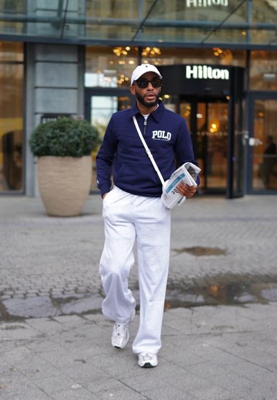 Un hombre viste un sudadera polo azul marino con el logo "Polo", pantalones deportivos blancos y zapatillas blancas. Sostiene un periódico, caminando fuera de un hotel Hilton.