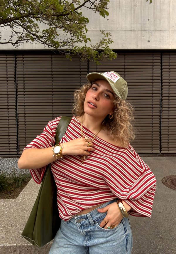 Jonge vrouw die een rood gestreepte off-shoulder top, blauwe spijkerbroek, groene pet en gouden sieraden draagt, poserend buiten tegen een bruine luikenachtergrond.
