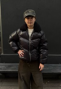 Black puffer jacket with a textured, soft collar; zip fastening; worn over a cream jumper and paired with dark green wide-leg trousers.