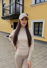 Conjunto de suéter de canalé beige y leggings a juego, con una gorra de color claro que lleva un logotipo, de pie frente a un fondo de edificio amarillo.
