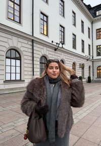 Brun konstpälsrock, grå scarf, mörka handskar och handväska med nyckelband. Har långt hår och står framför en vit byggnad.
