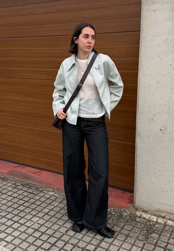 Kvinde med mørkt hår iført let jakke, hvid sweater, vide sorte bukser, sorte sko og crossbody-taske, stående ved en brun garagedør.