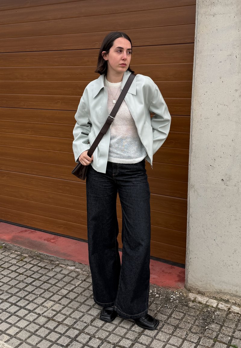 Femme aux cheveux foncés portant une veste claire, un pull blanc, un pantalon large noir, des chaussures noires et un sac bandoulière, debout près d'une porte de garage marron.