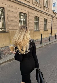 Žena kráčajúca po dlažobnej ulici, v ruke drží kávu v pohári so žltým slamkou. Má na sebe čierny priliehavý top a sukňu, nesie čiernu kabelku.