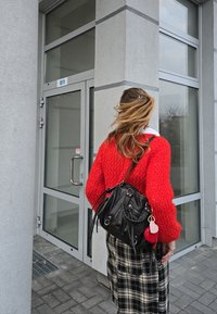 Heller roter, strukturierter Pullover kombiniert mit einer schwarzen Tasche mit Hardware-Akzenten, getragen zu einem karierter Rock. Grauer Gebäudeeingang im Hintergrund.