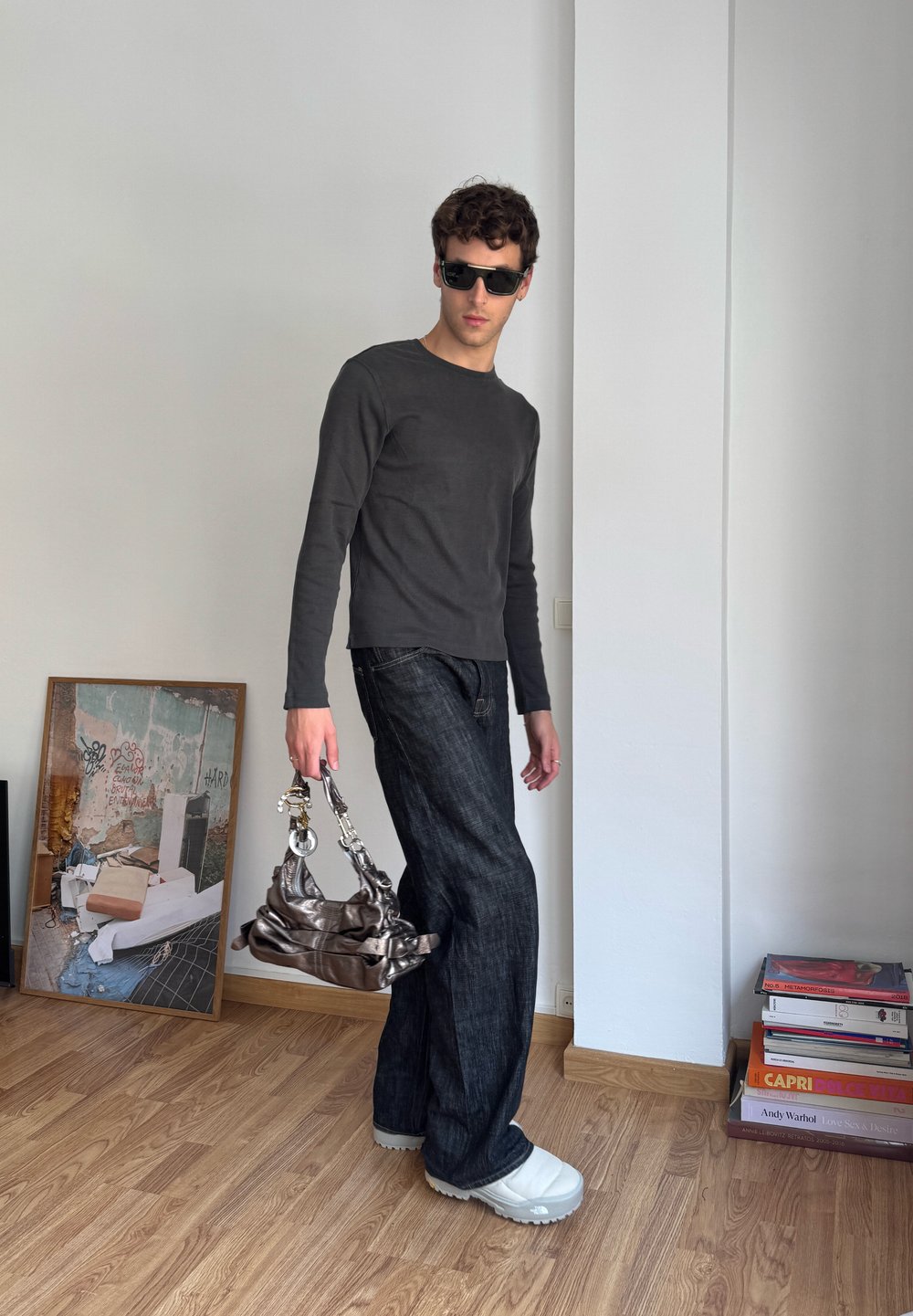 Dark long-sleeved shirt, black jeans, oversized light-coloured shoes, and a metallic bag. The model poses in a minimal indoor space.
