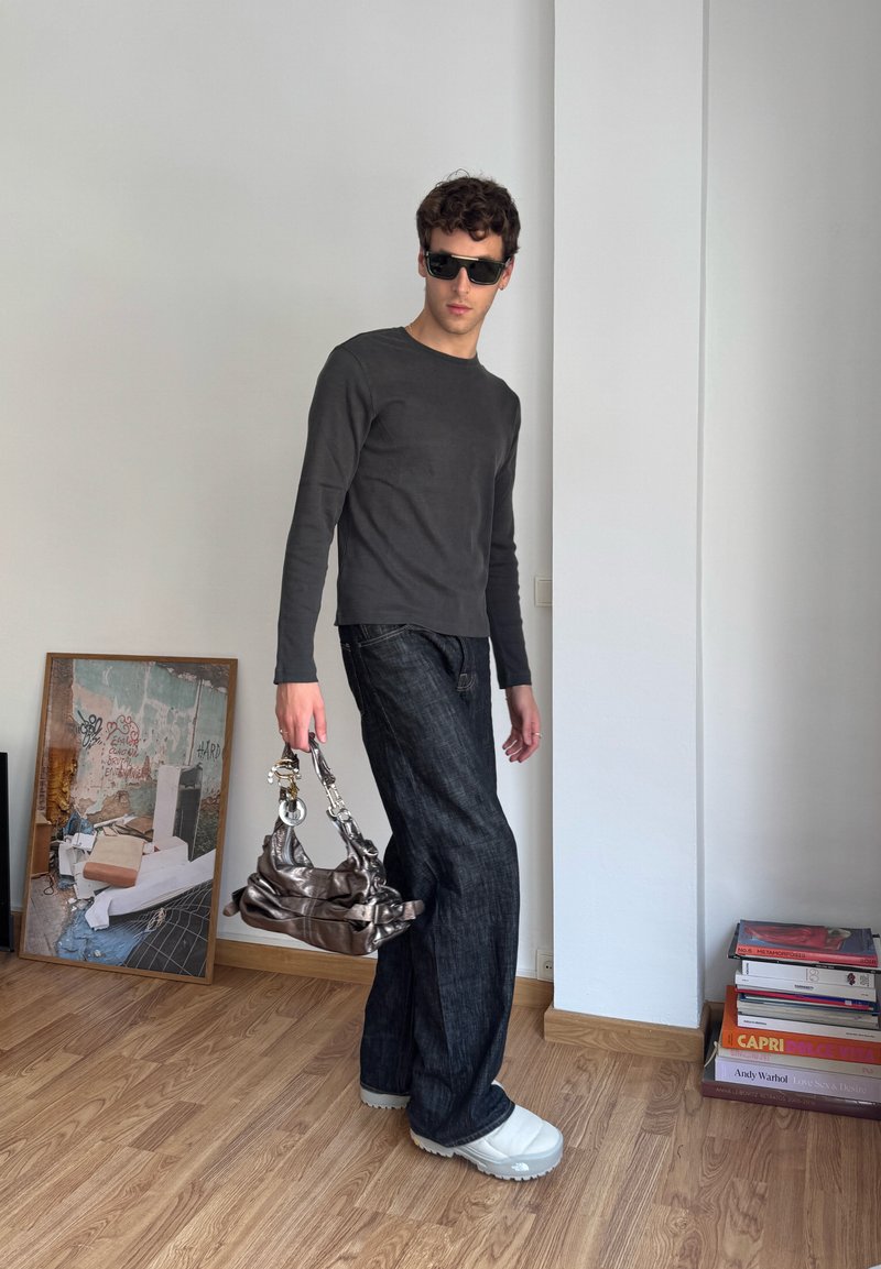 Maglietta scura a maniche lunghe, jeans neri, scarpe oversize di colore chiaro e borsa metallica. Modello in posa in uno spazio interno minimalista.