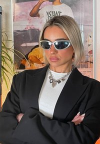 Črn blazer čez belo majico, srebrna masivna veriga s srčkastimi obeski in sončna očala s belim okvirjem. Kratki blond lasje in položaj s prekrižanimi rokami.