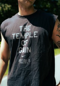 Camiseta preta sem mangas com o texto "O TEMPLO DO GAIN," logótipo da Nike e "FAZ. DESDE 1972" em branco. Textura de tecido suave e desgastado.