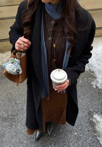 Femme portant un manteau noir, un pull marron et une jupe, tenant un gobelet de café à emporter et un sac à main marron avec un porte-clés en peluche gris.