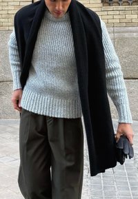 Pull gris clair côtelé avec un col montant, associé à un pantalon ample vert foncé et une écharpe noire drapée sur les épaules. Gants noirs en main.