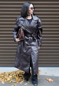 Brauner Leder-Trenchcoat mit tailliertem Gürtel, Reverskragen und Schlitzsaum, kombiniert mit einer kleinen braunen Handtasche und schwarzer Hose.