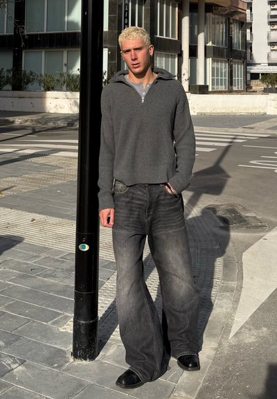 Suéter gris de canalé con cuello de cremallera, combinado con jeans de pierna ancha en negro desgastado y zapatos negros, de pie cerca de un poste de calle sobre el pavimento.