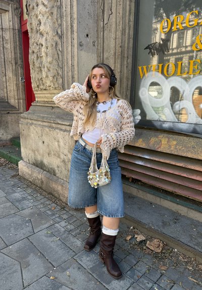 Cardigan de ganchillo beige sobre una blusa blanca, falda de mezclilla azul, botas marrones hasta la rodilla y un bolso de cuentas; usando auriculares negros.