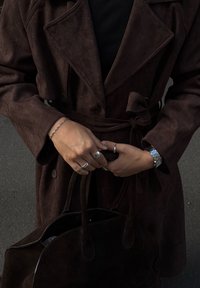 Sötétbarna kabátot viselő személy, aki a övet köti, sötétbarna táska a kezében, ezüst gyűrűk, karkötő és óra van rajta sötét háttér előtt.