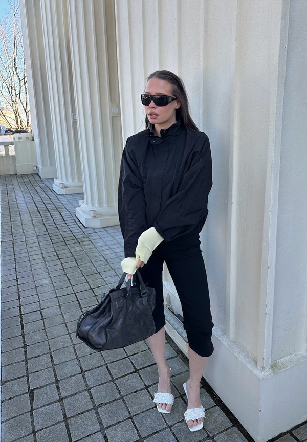 Nainen mustassa takissa ja polvipituisissa housuissa, aurinkolasit päässään, pitää suurta mustaa käsilaukkua, seisoo valkoisen pylväikkörakennuksen vieressä.