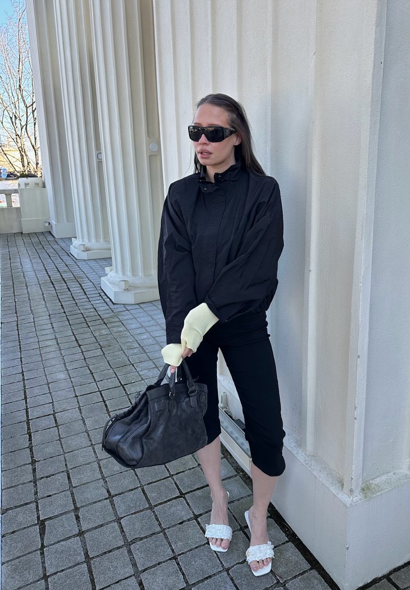 Frau in schwarzer Jacke und gekürzten Hosen mit Sonnenbrille, hält eine große schwarze Handtasche, steht bei einem weißen Gebäude mit Säulen.