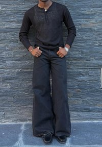 Camisa de manga larga negra con textura acanalada y botón en la placket, combinada con pantalones de mezclilla anchos negros y zapatos negros, contra un fondo de pared de piedra.