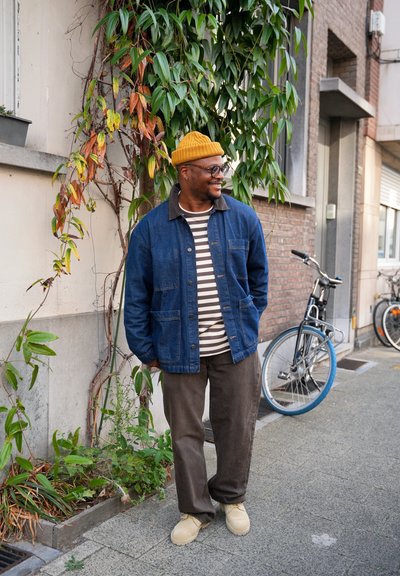 Chaqueta de mezclilla, camisa a rayas, pantalones marrones y gorro de punto amarillo. Textura suave, diseño casual. Follaje verde y bicicletas de fondo.