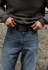 En person som bär en grå stickad tröja och blå Levi's jeans justerar ett svart bälte med ett metallspänne från Levi Strauss & Co.