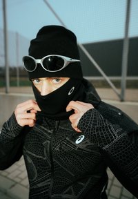 Veste noire à fermeture éclair avec un motif géométrique, portée avec un bonnet noir et un masque facial. Des lunettes de soleil réfléchissantes sont posées sur le dessus.