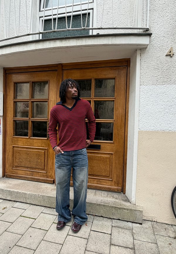 Uomo con dreadlocks, indossa un maglione a righe rosse e nere e jeans larghi, in piedi davanti a una porta di legno con pannelli in vetro su un marciapiede in pietra.