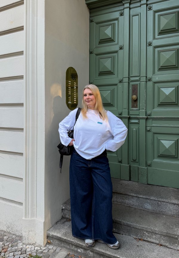 Femme aux longs cheveux blonds portant une chemise blanche à manches longues et un jean bleu large, debout sur des marches près d'une grande porte verte et de la façade d'un bâtiment.