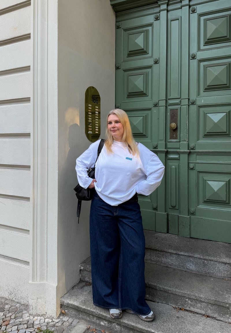 Woman with long blonde hair wearing white long-sleeve shirt and wide blue jeans stands on steps by large green door and building facade.