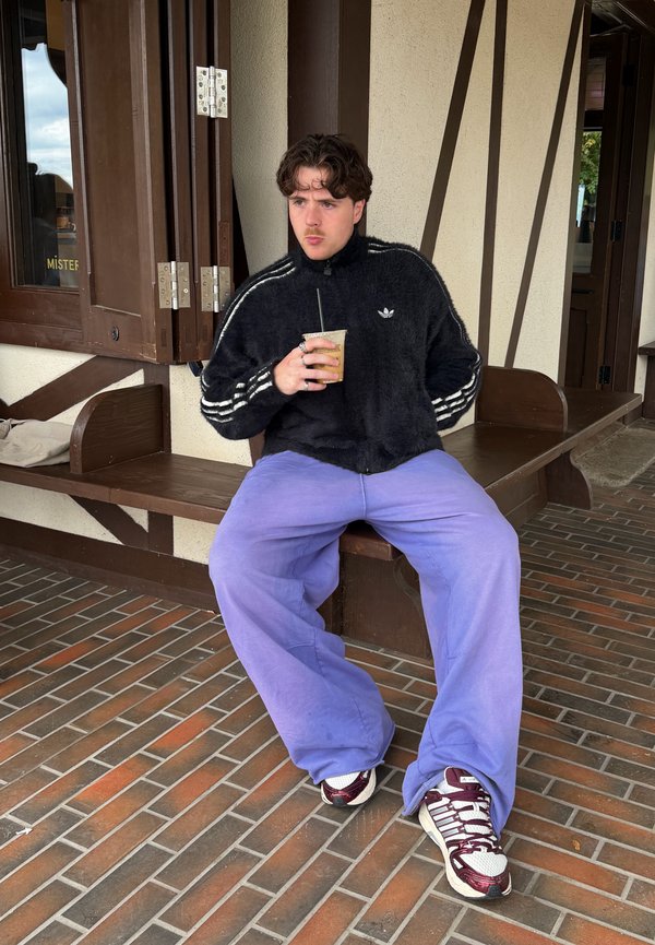 Jeune homme en veste Adidas noire et pantalon violet assis sur un banc en bois en train de boire un café glacé en plein air.