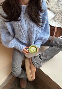 Femme portant un pull bleu en tricot et un pantalon rayé, assise les jambes croisées, tenant une tasse de matcha latte vert avec un dessin en latte art.