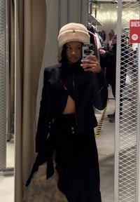 Μαύρο cropped μπουφάν με φερμουάρ, τοποθετημένο πάνω από μια μαύρη μπλούζα και φούστα. Μπεζ γούνινο καπέλο. Η αντανάκλαση στον καθρέφτη δείχνει το background ενός καταστήματος ρούχων.