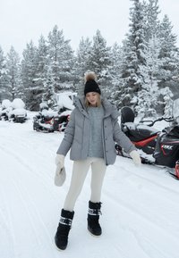 Giubbotto puffa grigio, maglione grigio chiaro, leggings crema e stivali da neve neri. Tiene dei guanti crema, paesaggio innevato con motoslitte.