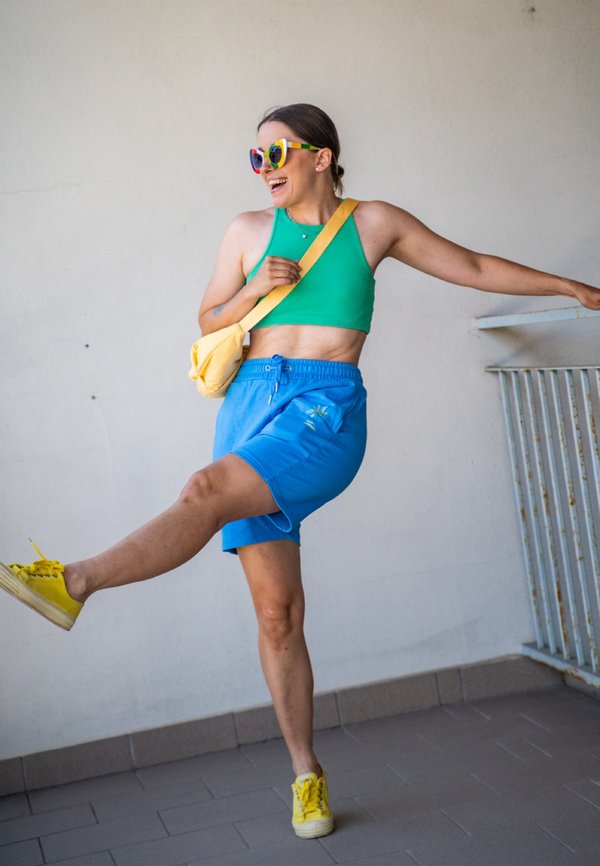 Kvinde i farverige solbriller, grøn cropped top, blå shorts og gule sko, der sparker benet op, mens hun smiler på et flisebelagt gulv nær gelænder.