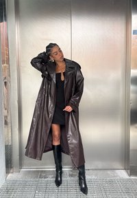 Lång brun trenchcoat i konstläder med bred krage och dubbelknäppt knappslå, kombinerad med en figurnära svart klänning och knähöga stövlar.