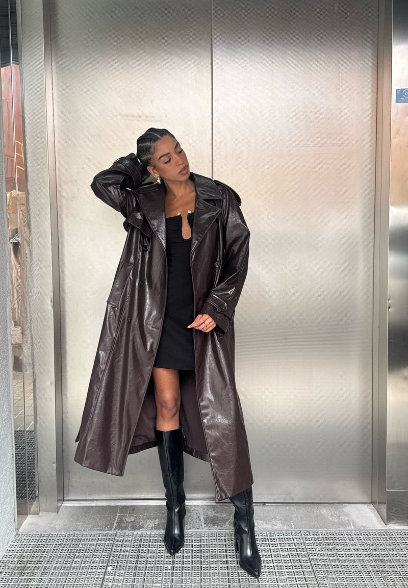 Ilgas, rudos spalvos dirbtinės odos tranšėjos paltas su plačia apykakle ir dvigubais sagomis, derinamas su aptempiama juoda suknele ir ilgaauliais iki kelių.