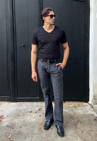 Zwarte v-hals t-shirt, gestreepte grijze broek, zwarte riem en glanzende zwarte schoenen. Zonnebril op; staand tegen een achtergrond van een zwarte deur.