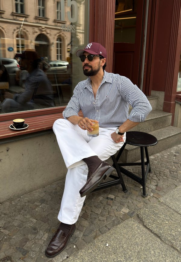 Homme portant une chemise rayée et un pantalon blanc assis à l'extérieur d'un café sur un tabouret noir, tenant un verre avec une boisson, à côté d'une tasse de café.
